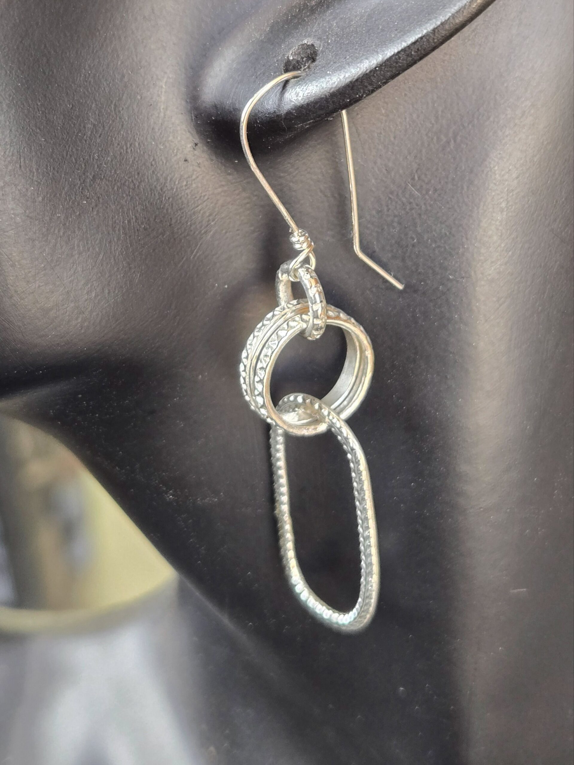 Textured Link Sterling Silver Earrings. French wire, attached to a loop, attached to two textured round rings, attached to one long textured oval shown on a mannequin to see the drop length.